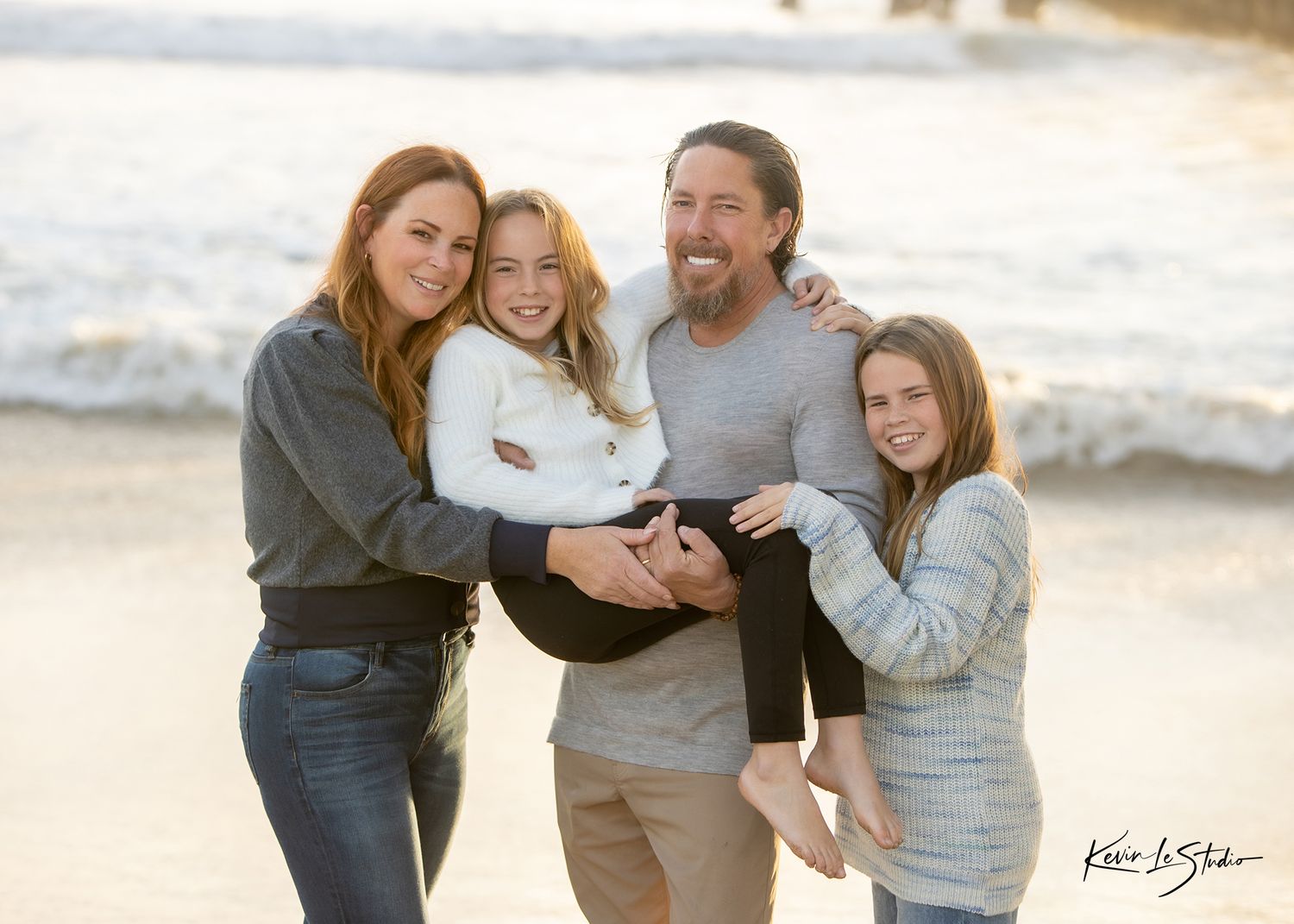 Happy family of four laughing together on a sunny beach, parents holding two girls in a warm outdoor portrait.