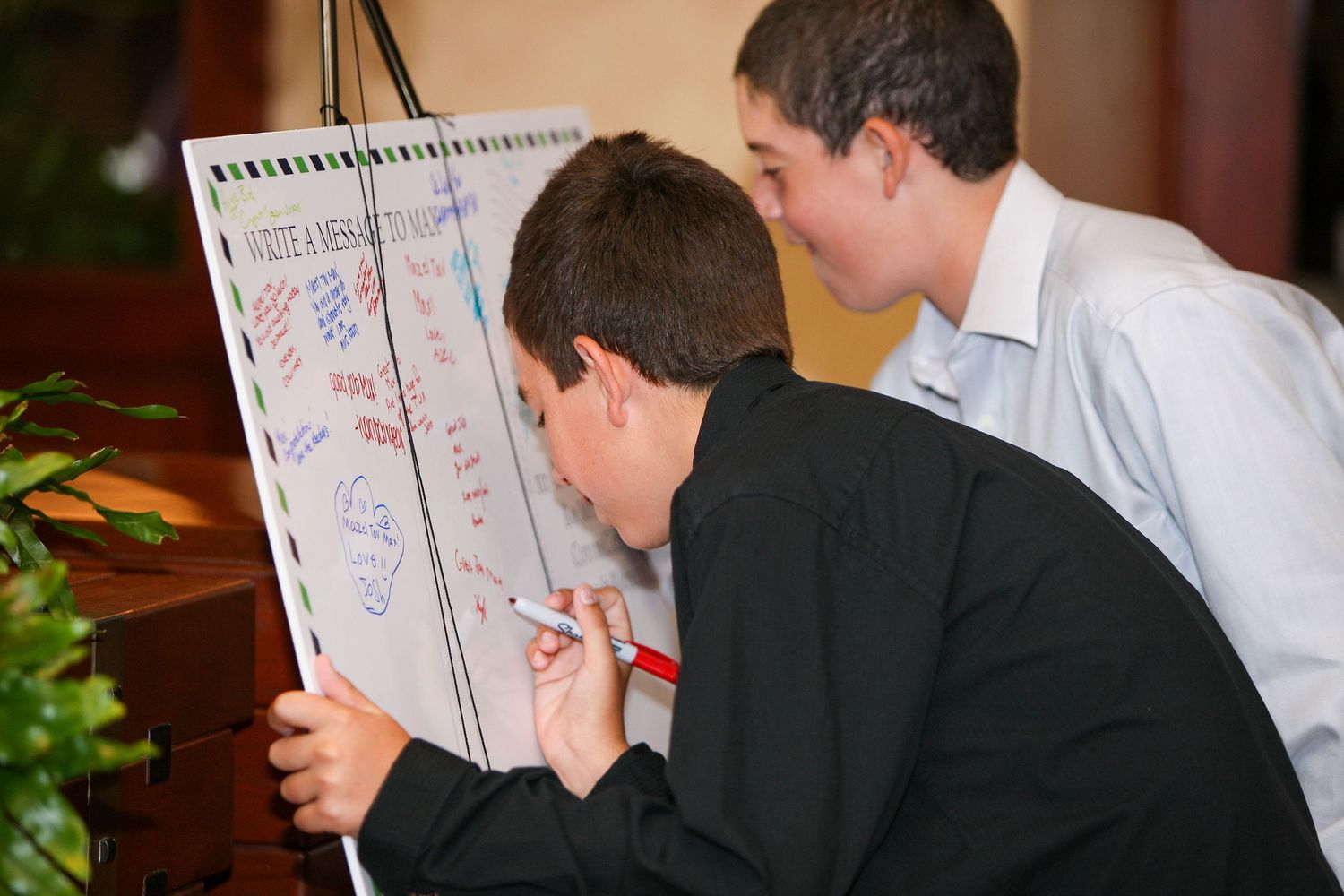 Two boys in formal attire signing a large whiteboard display at an event or celebration.