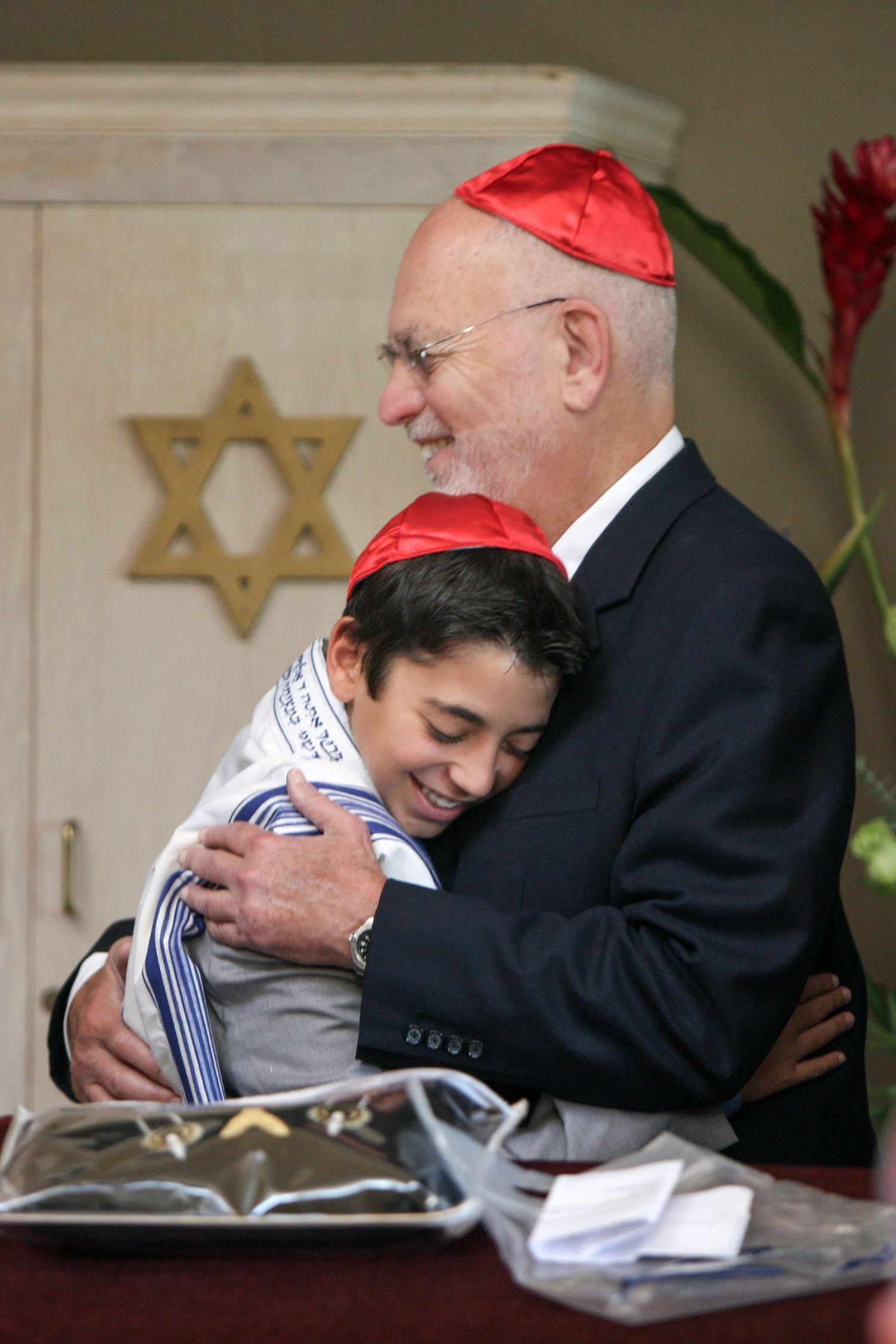 Elderly man and young boy sharing a warm hug at a Bar Mitzvah ceremony, both wearing red kippot.