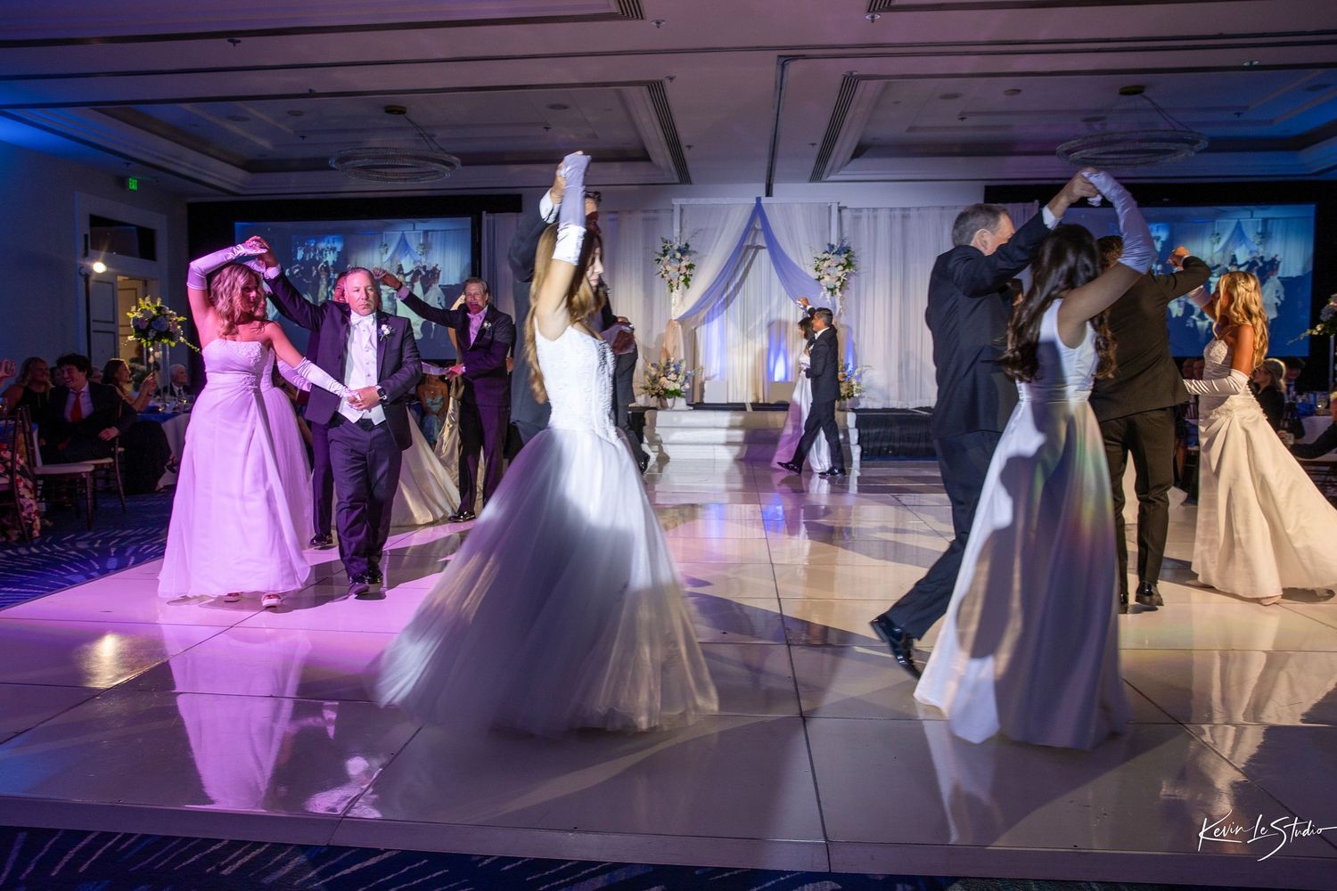 Elegant wedding reception with bride and groom dancing on white floor under purple lights in a decorated ballroom.