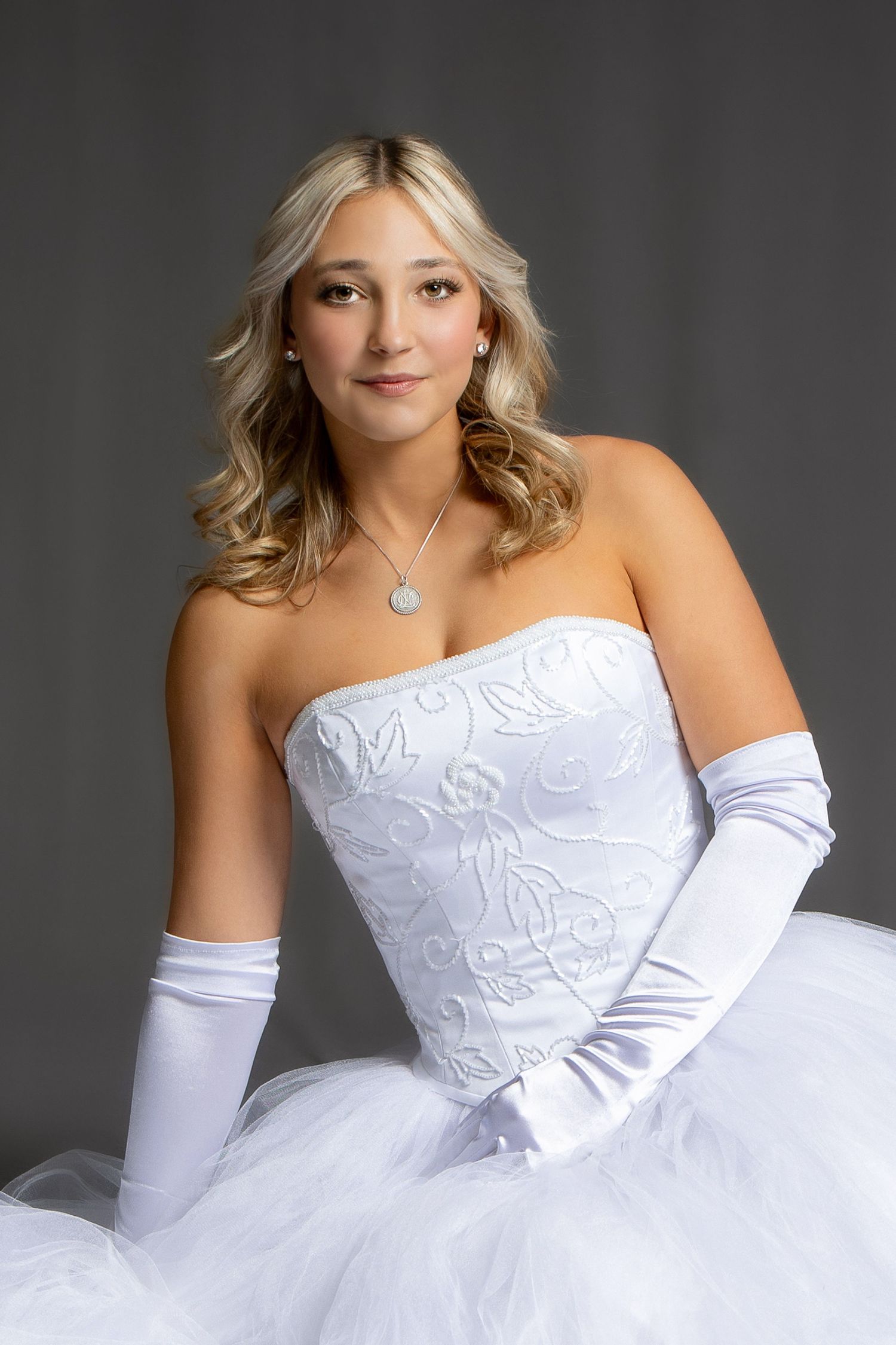 Young woman in white strapless ballgown with satin gloves and pendant necklace poses elegantly against gray background.