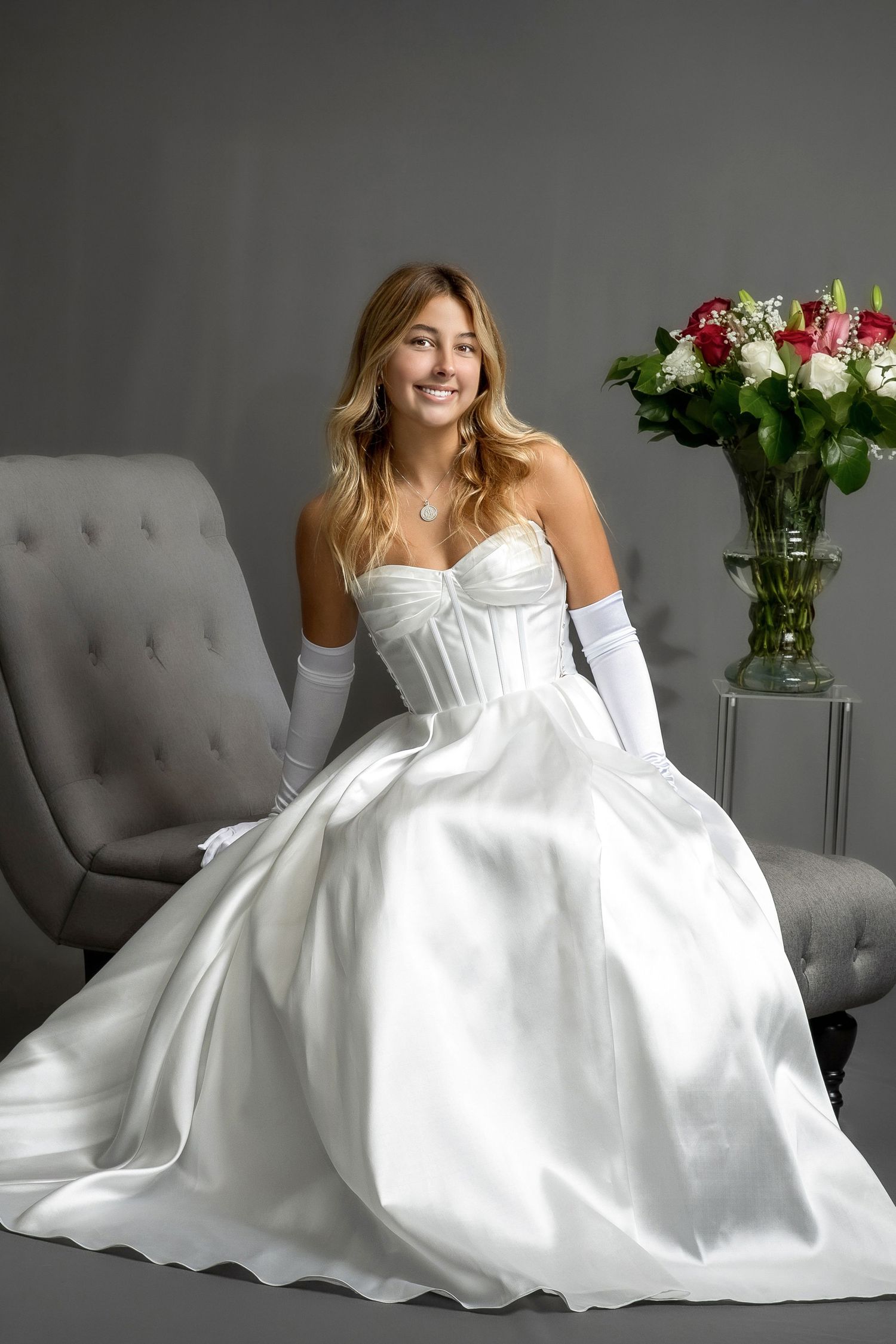A smiling bride in a white satin gown and gloves sits on a gray chair beside a vase of red and white roses.
