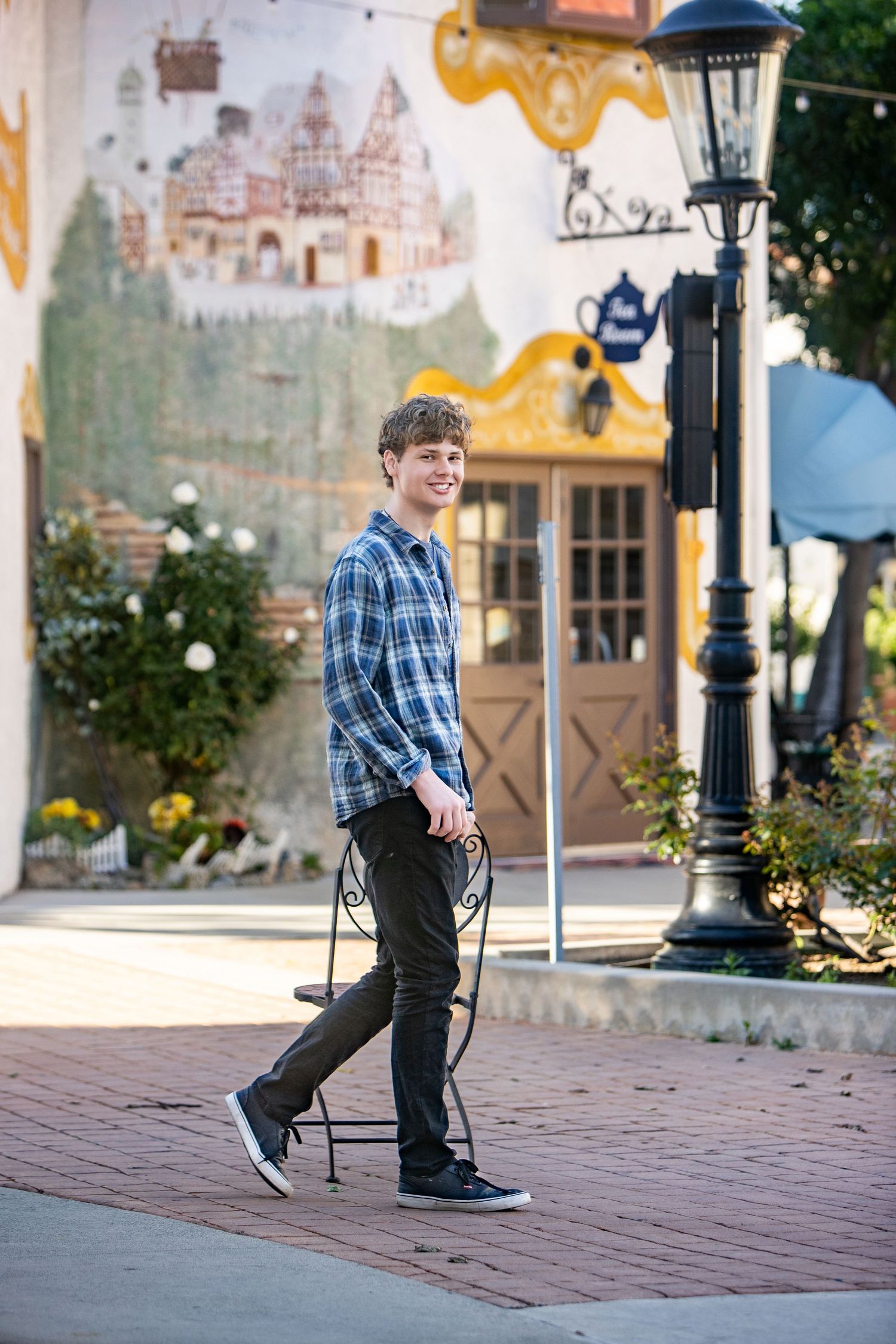 Smiling teen boy in plaid shirt and black jeans walks on brick plaza beside ornate lamp post with colorful mural behind.