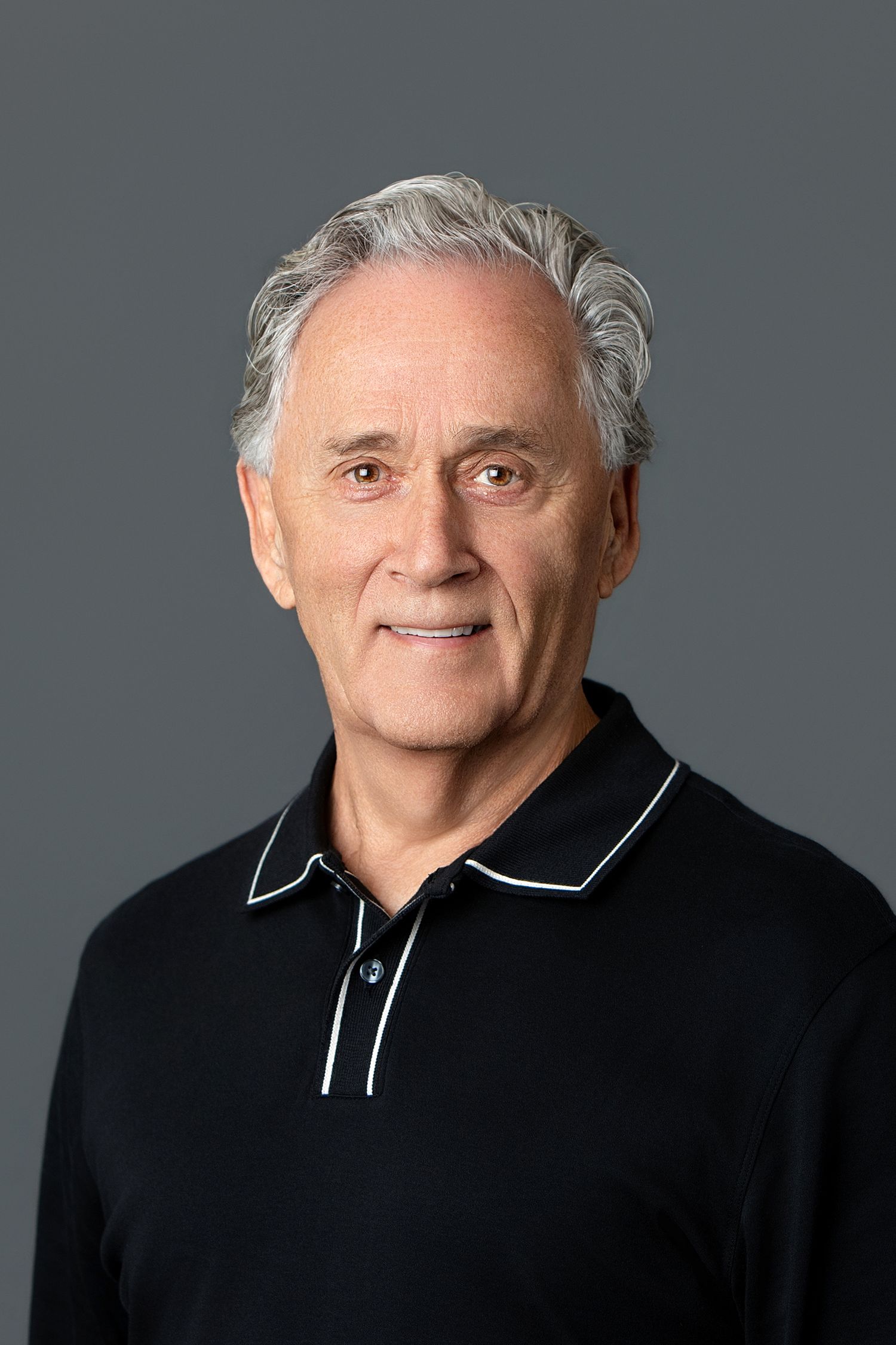 Smiling older man with gray hair wearing a black polo shirt, photographed against a gray studio background.
