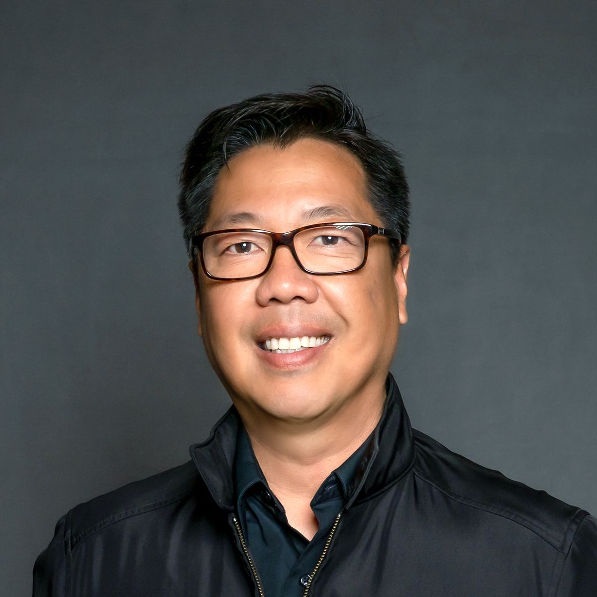 Professional headshot of a smiling middle-aged Asian man wearing black-rimmed glasses and a black jacket against a gray background.