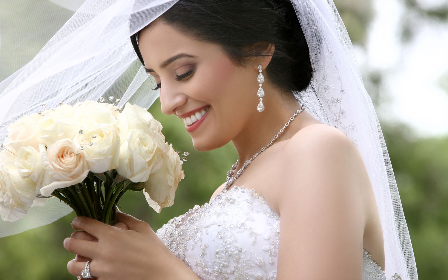 Beautiful bride smiling while holding white rose bouquet wearing lace gown and pearl jewelry.
