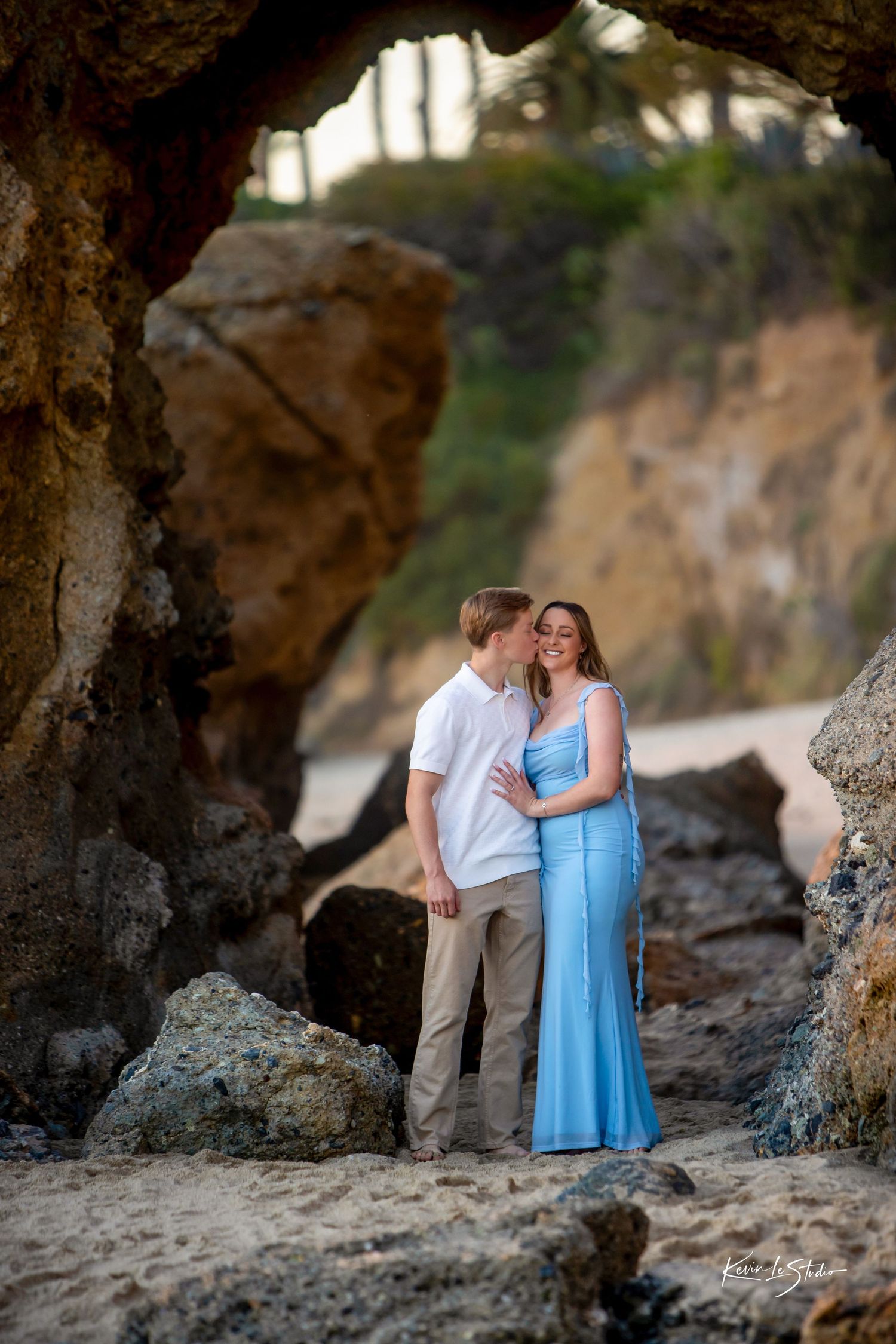 Couple sharing a kiss framed by a natural rock arch on a sandy beach at sunset.