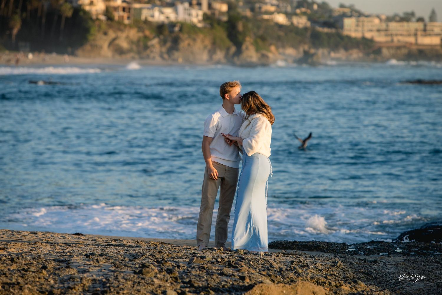 Couple kissing on rocky beach shore with coastal cliffs in background.