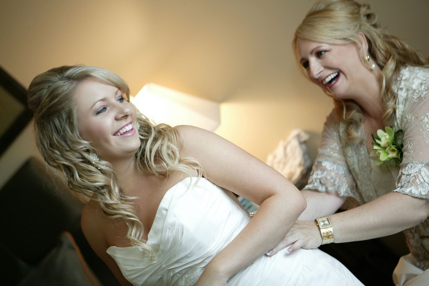Two blonde women laughing joyfully, bride in white lace wedding dress being helped by older woman.