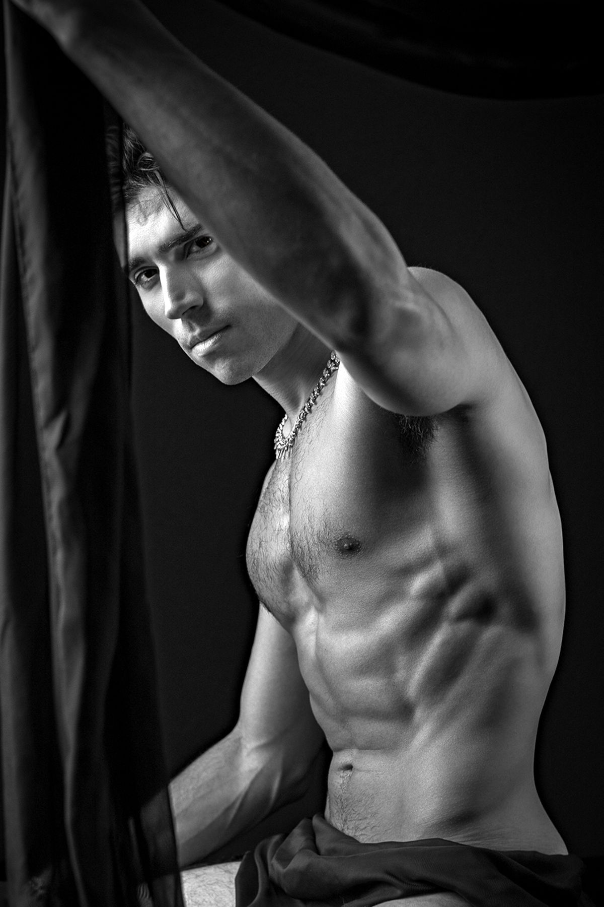 Black and white photo of a muscular shirtless man posing dramatically beside dark fabric draping.