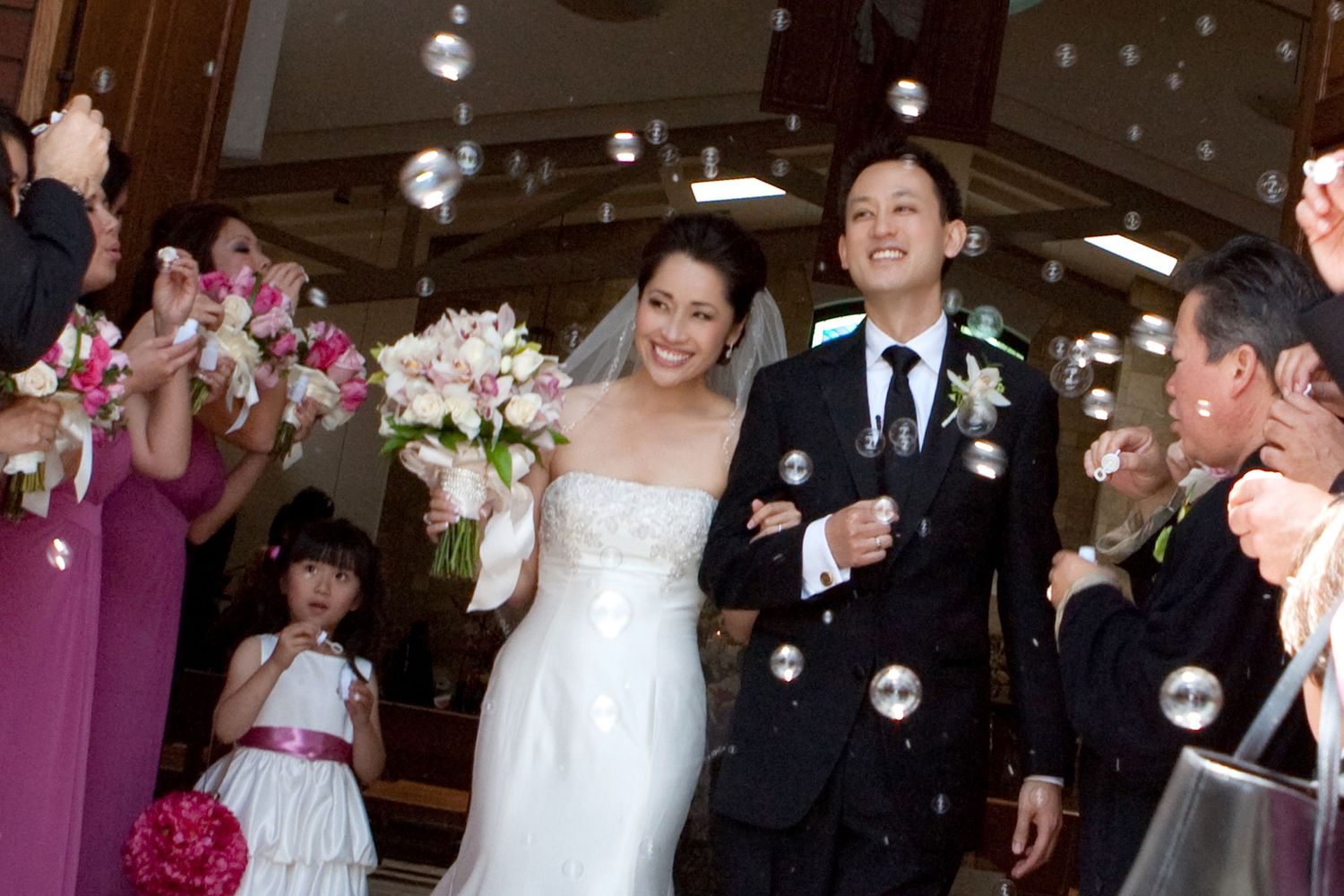 Newlywed Asian couple exits church amid bubbles, bride in white gown holding bouquet, groom in black suit, guests celebrating.