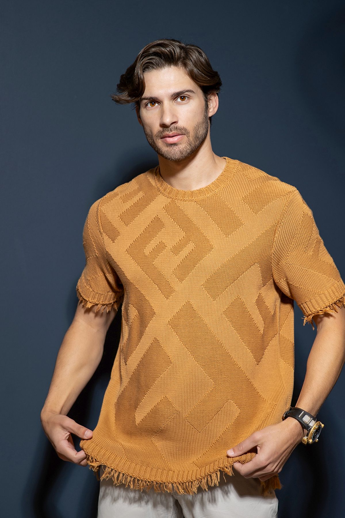 Man wearing an orange geometric patterned knit t-shirt with fringe hem, posing against a dark background.