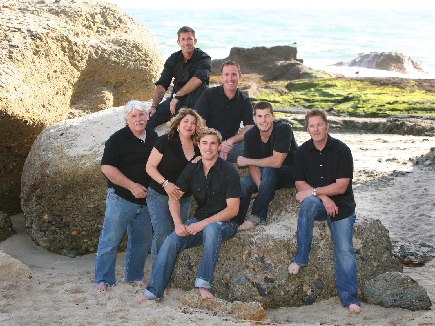 Group of six adults in matching black shirts posing on rocky coastal boulders at the beach.