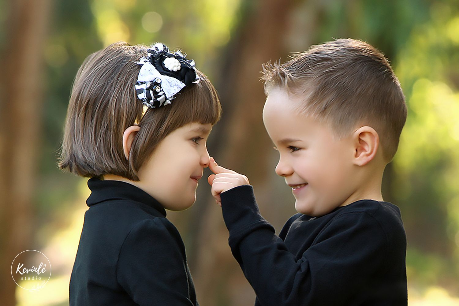 Two young children facing each other as a boy gently touches a girls nose in an outdoor park setting.