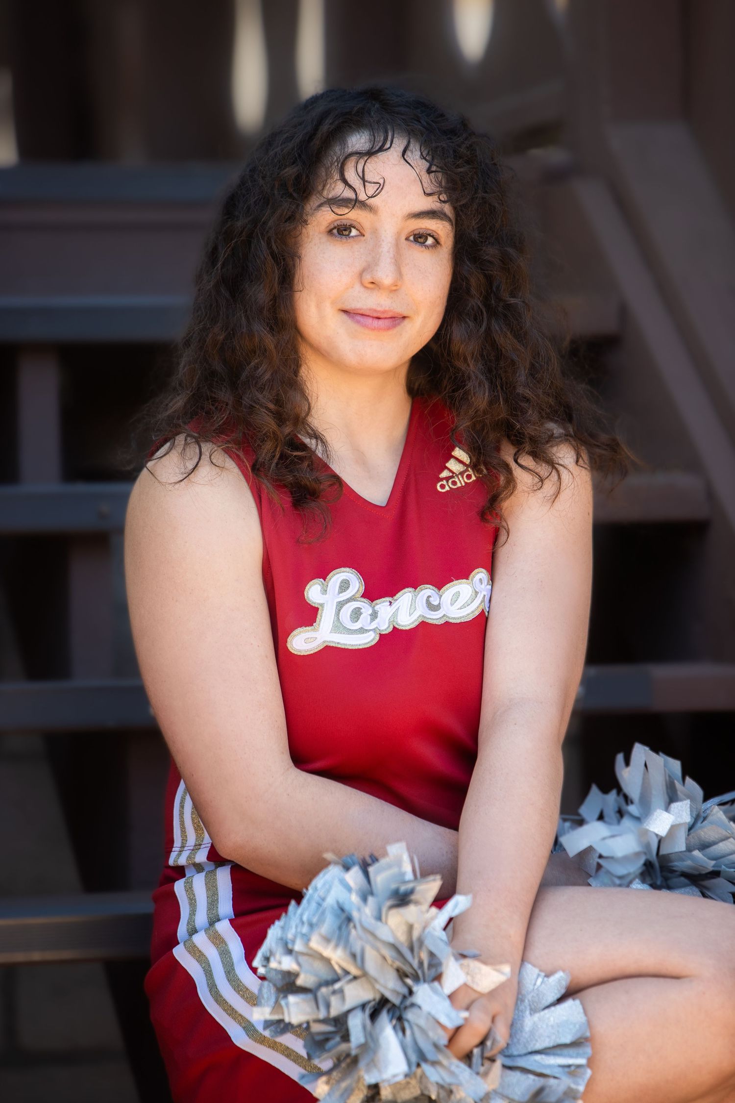 Young cheerleader in red Lancers uniform sits on bleachers holding silver pom-poms.