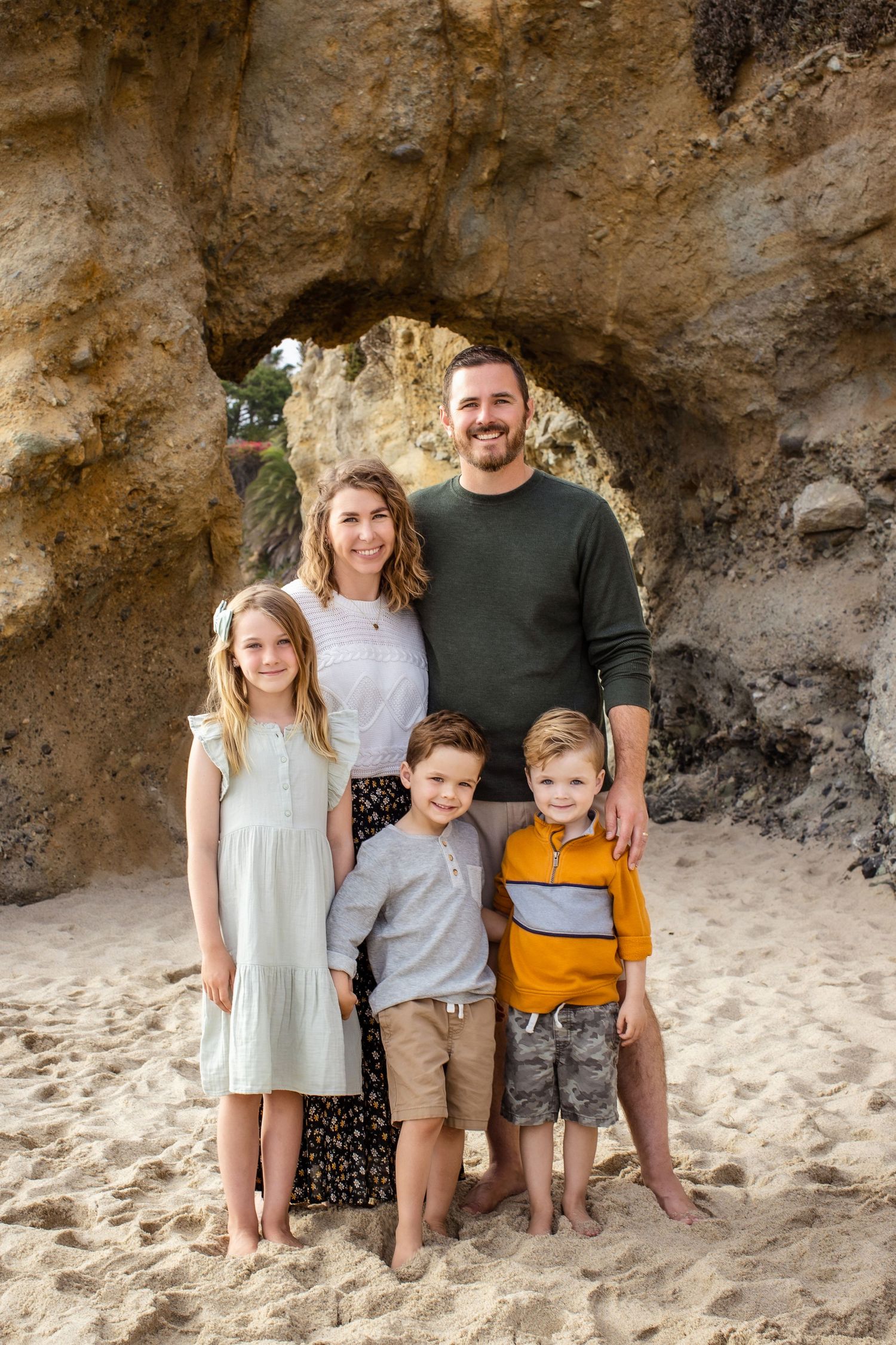 Happy family of five posing under natural rock arch on sandy beach during photo session.