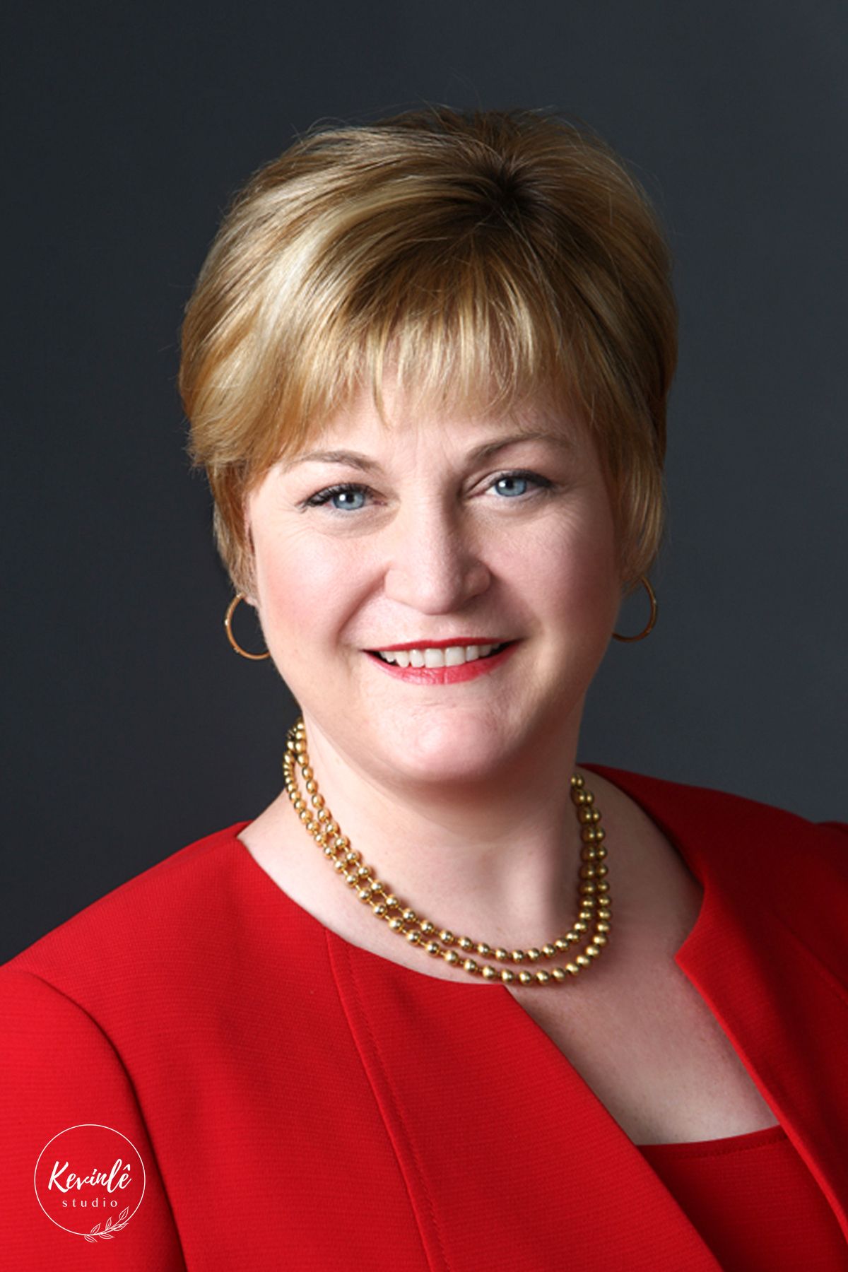 Professional headshot of a smiling blonde woman in a red blazer with a gold pearl necklace on gray background.