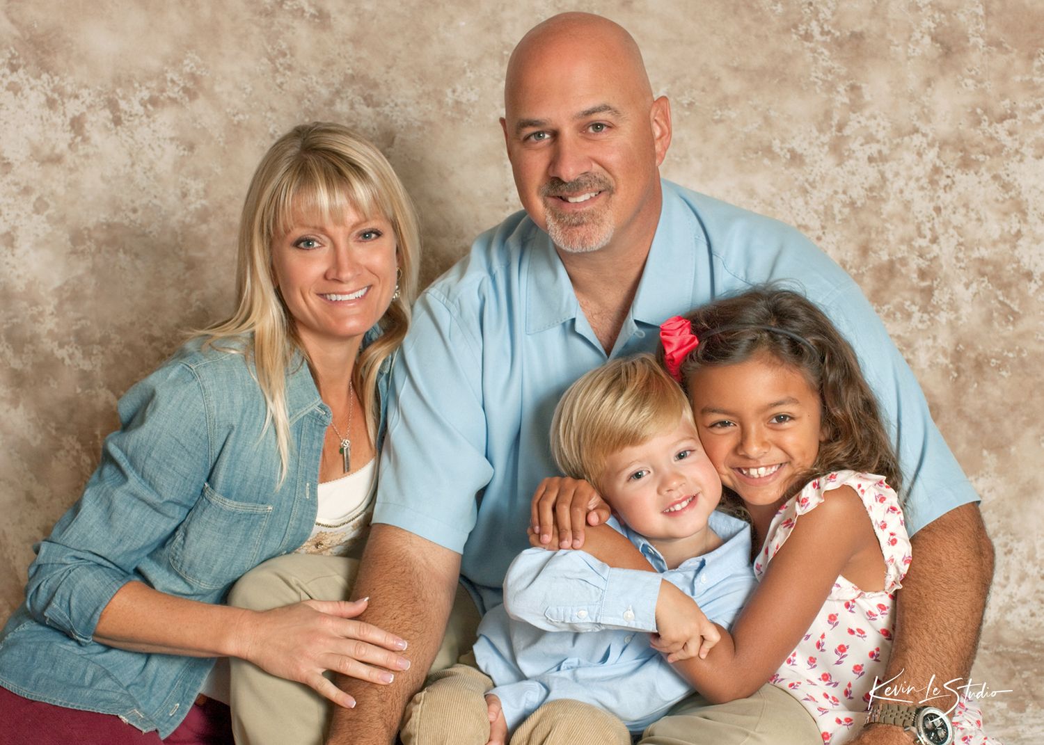 Family of four posing for a studio portrait, with parents and two young children smiling warmly against a beige background.