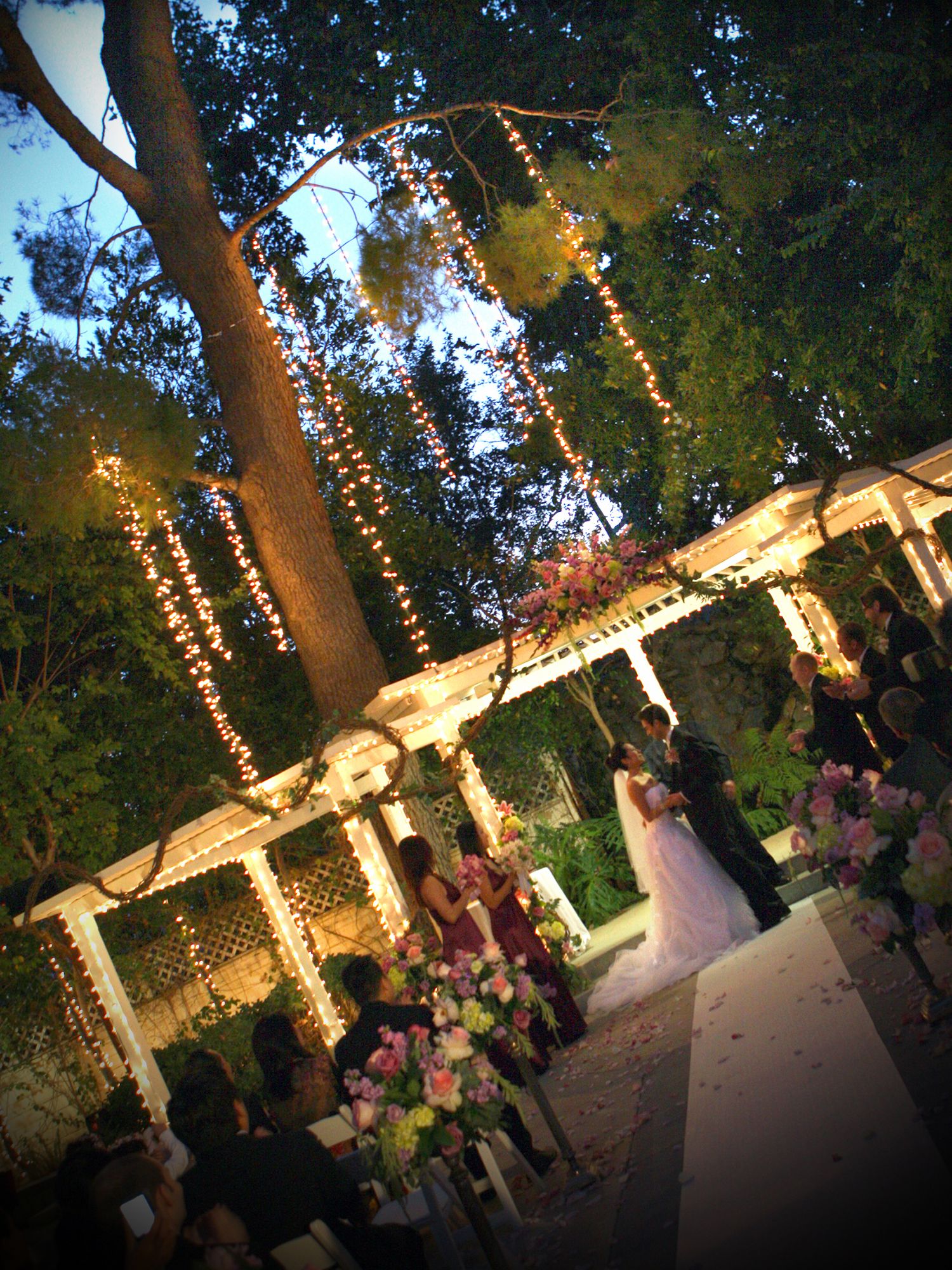 Romantic outdoor wedding ceremony at night under twinkling fairy lights and a white pergola surrounded by lush trees.