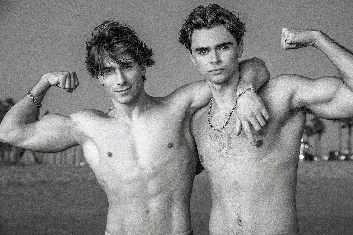 Two shirtless muscular young men flexing biceps together in a black and white outdoor photo.