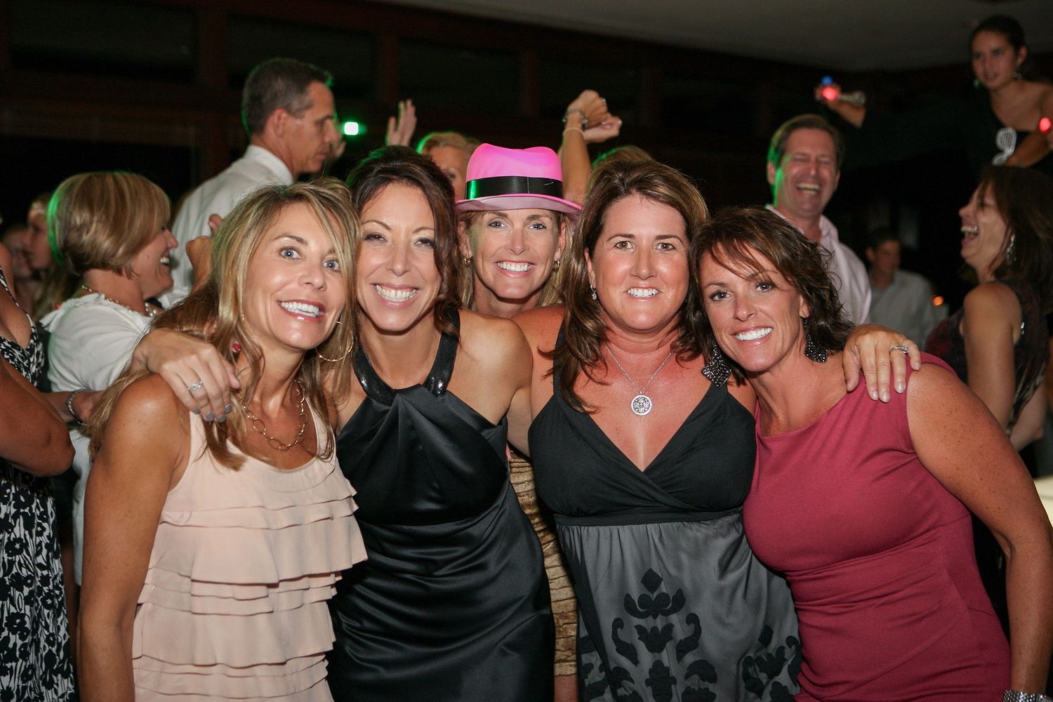 Five women posing together at a party, one wearing a pink hat, smiling at the camera.