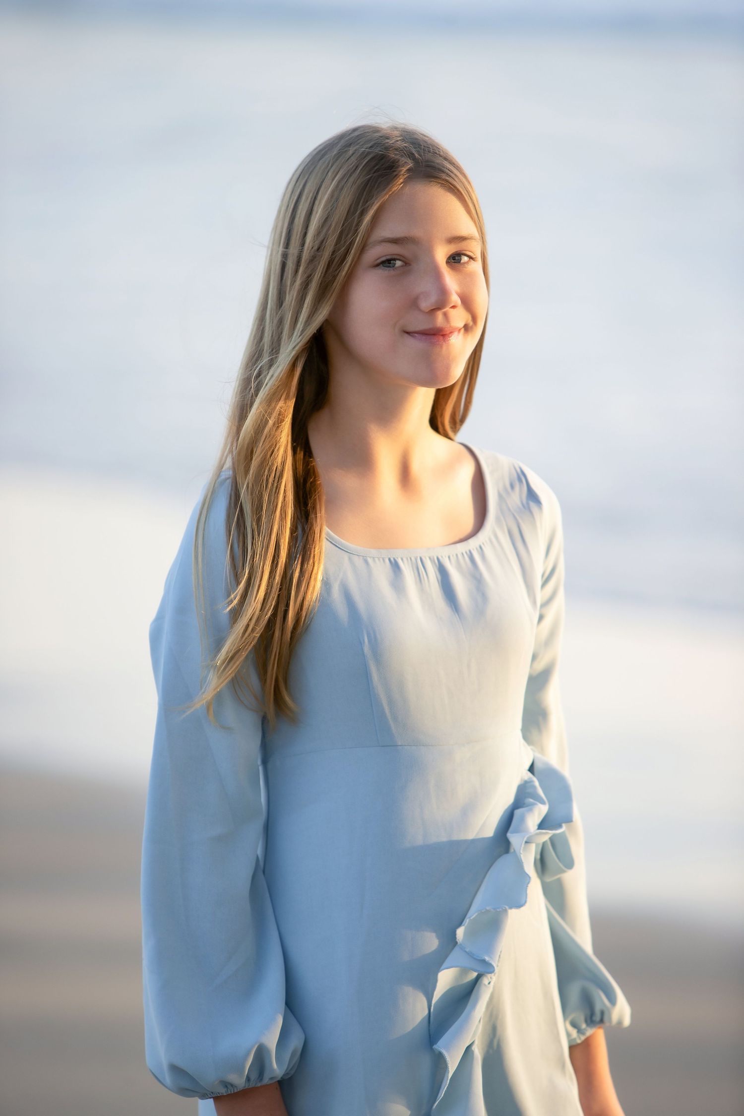 Young blonde girl in a light blue dress smiling at the beach during golden hour sunset.