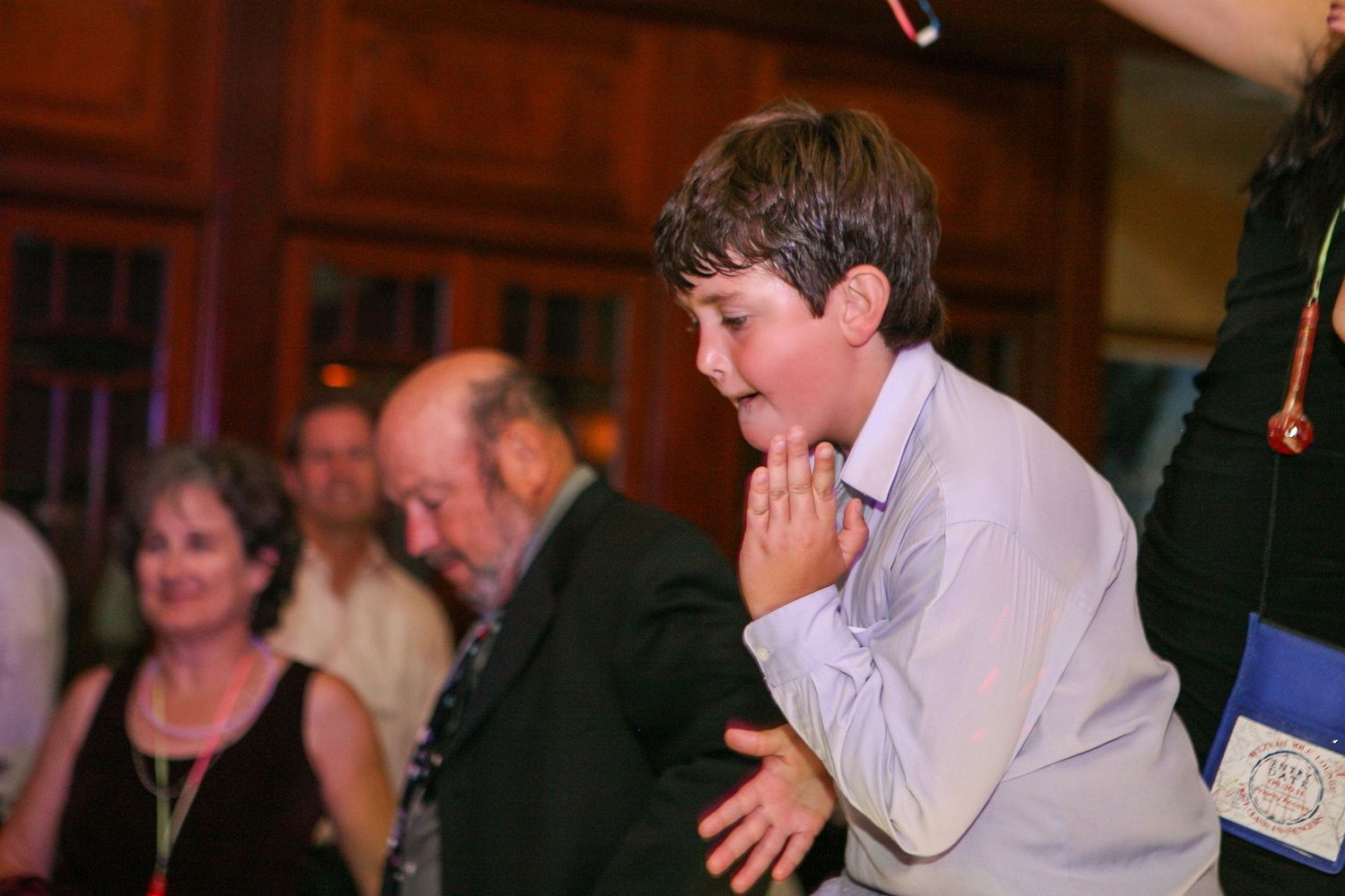 Excited young boy with hands clasped at an indoor event, with adults socializing in the background.