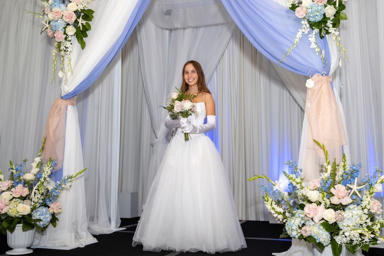 Bride in white ballgown holding bouquet stands under blue and white draped floral arch at elegant wedding ceremony.