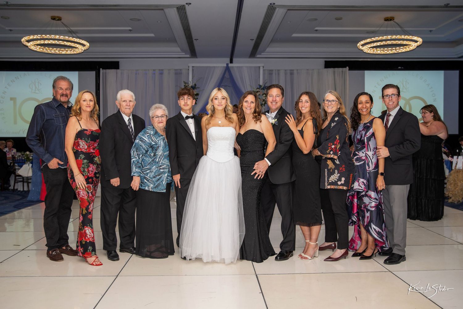 A large family group poses together in formal attire at an elegant ballroom event with chandeliers and screens in the background.