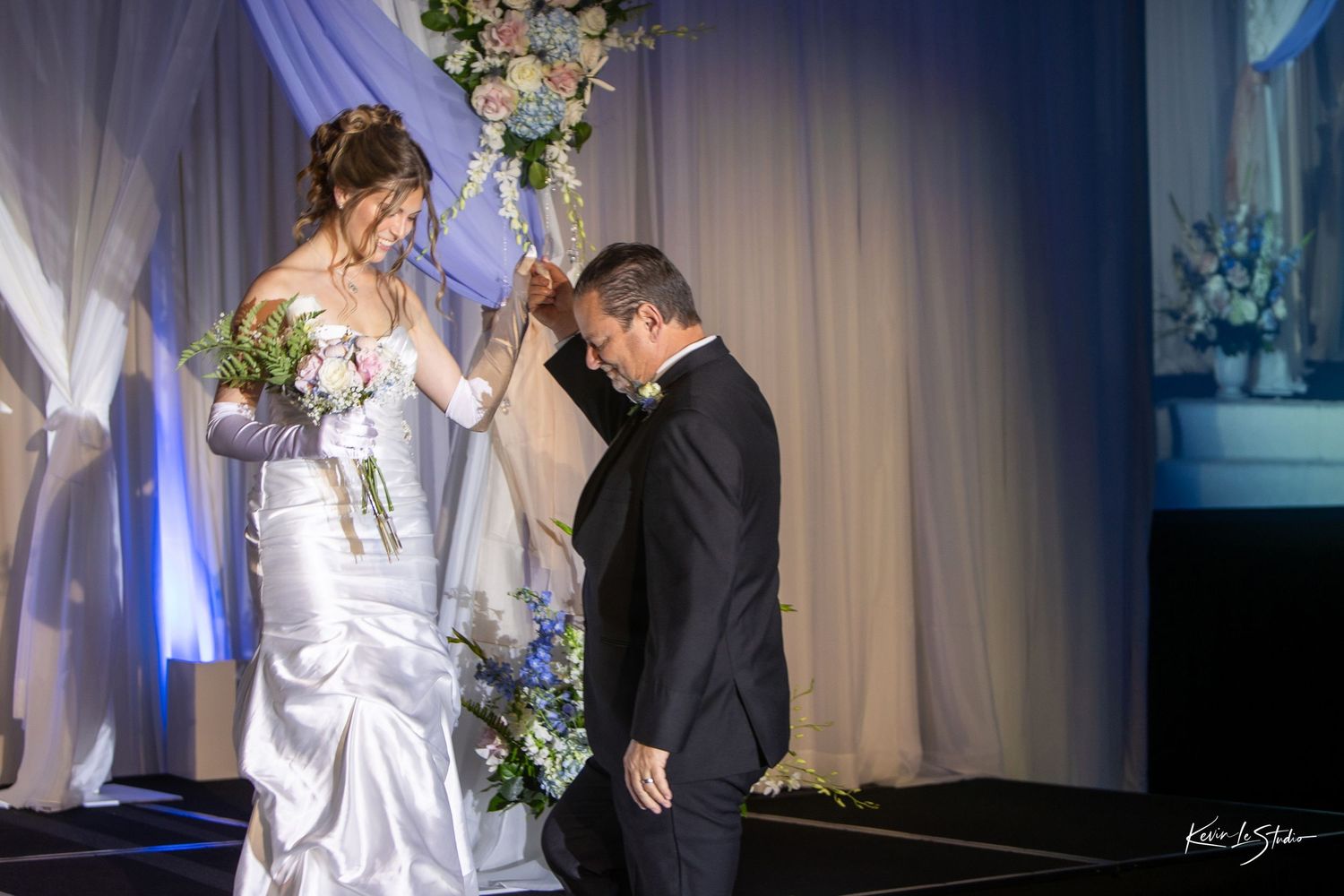 Bride in white gown places hand on groom's bowed head during elegant indoor wedding ceremony.