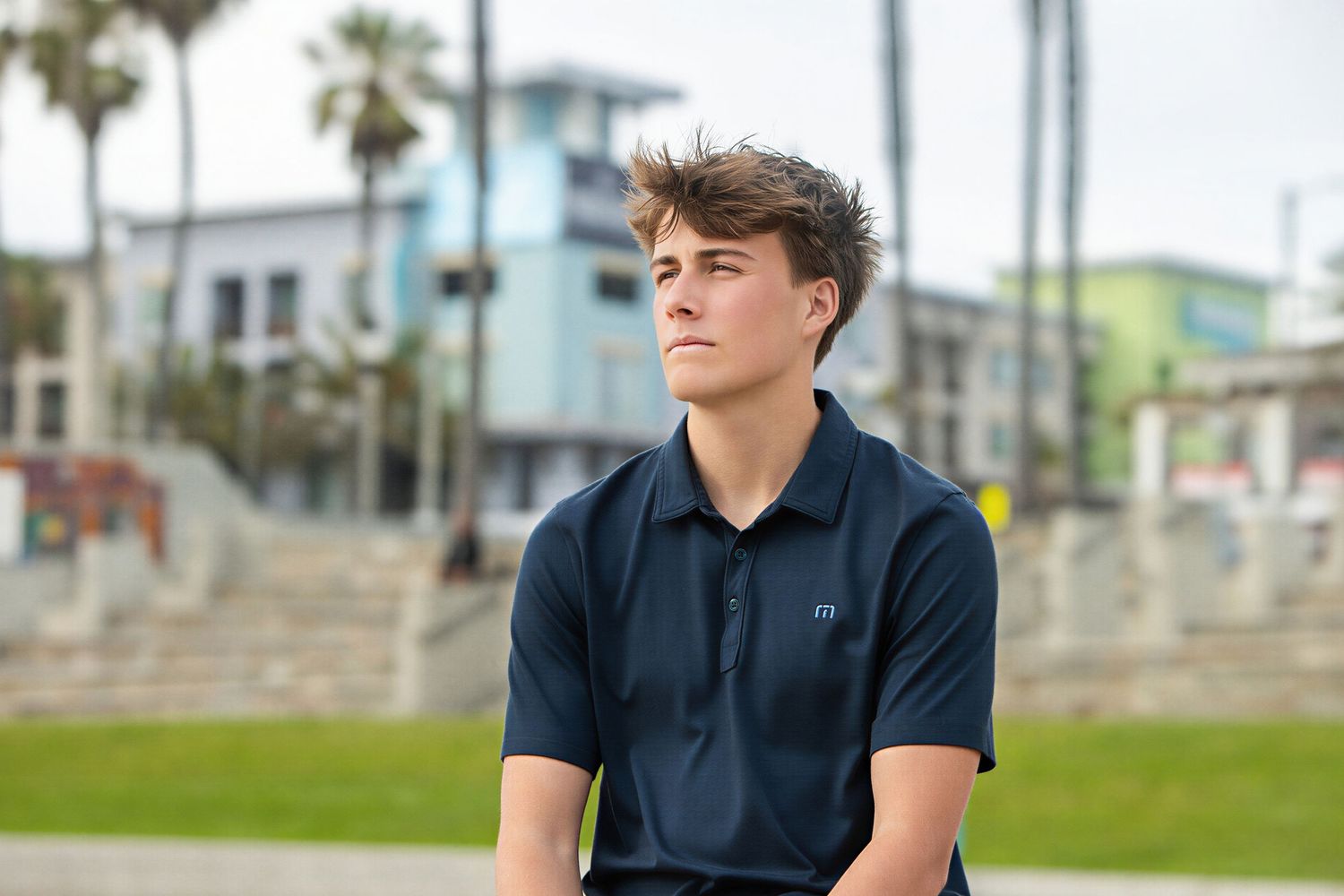 Young man in navy polo shirt sitting outdoors near palm trees at Venice Beach on a sunny day.