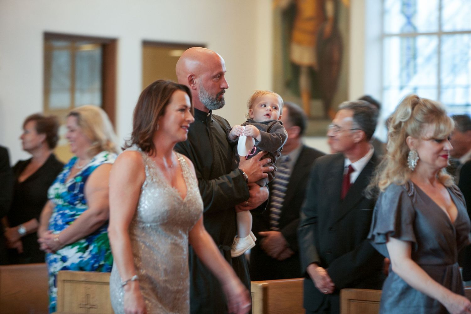 Family with baby attending a church ceremony, woman in elegant dress standing beside bald man holding infant.