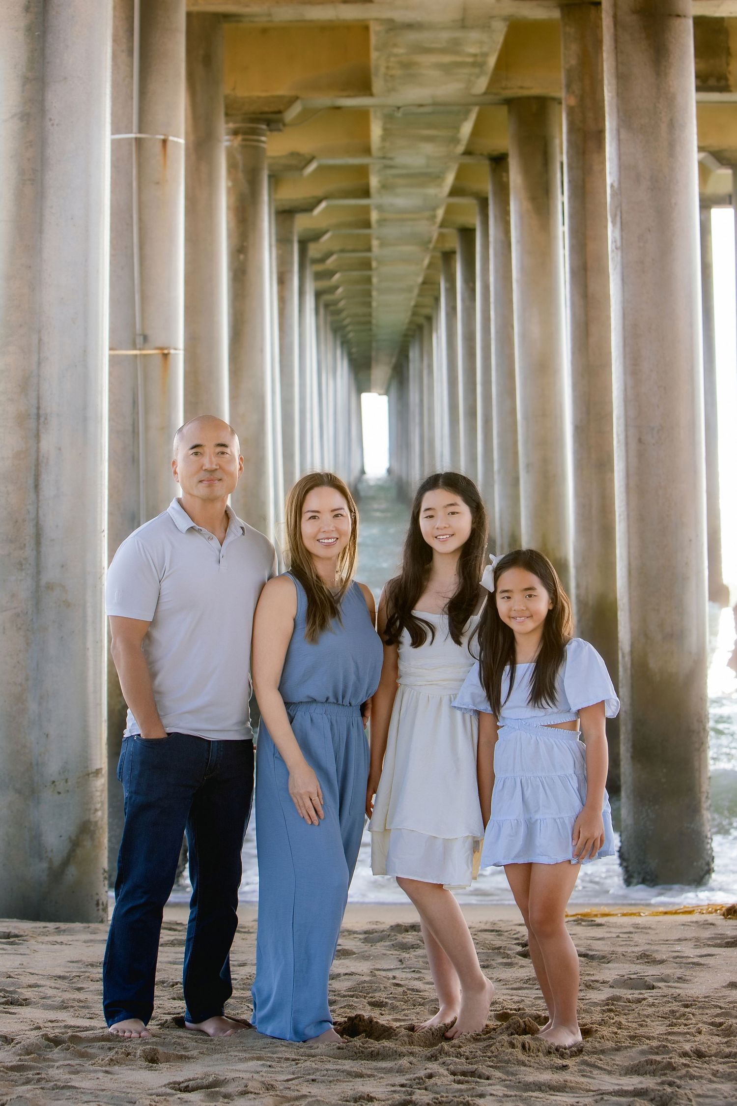 Family of four smiling under a beach pier on sandy shore, wearing casual blue and white outfits.