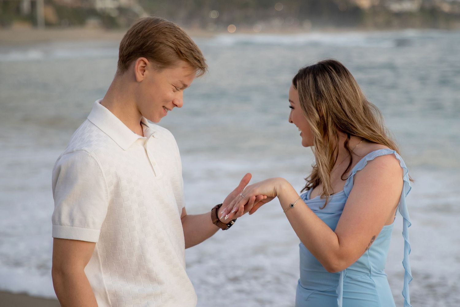 Young couple on beach examining engagement ring in soft golden light.