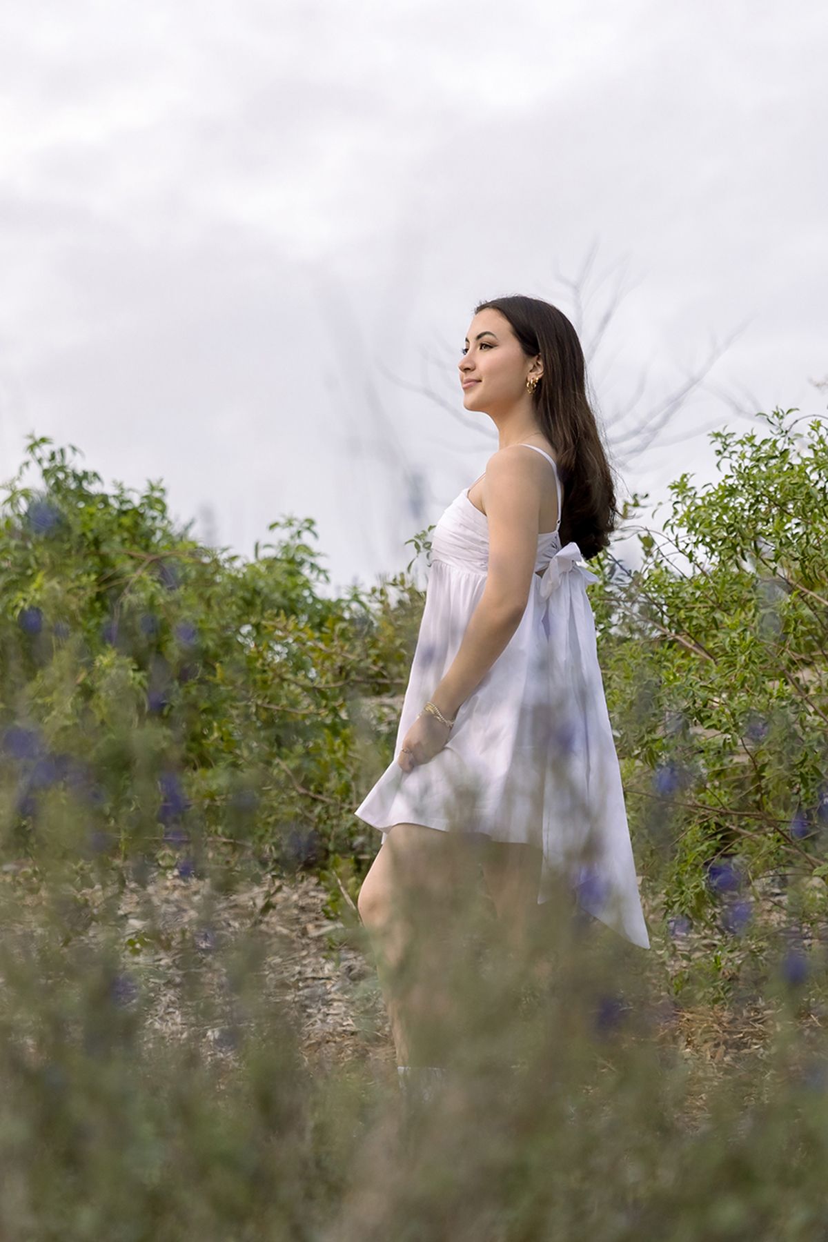 Young woman in flowing white dress stands among wildflowers and blue blooms under cloudy sky.