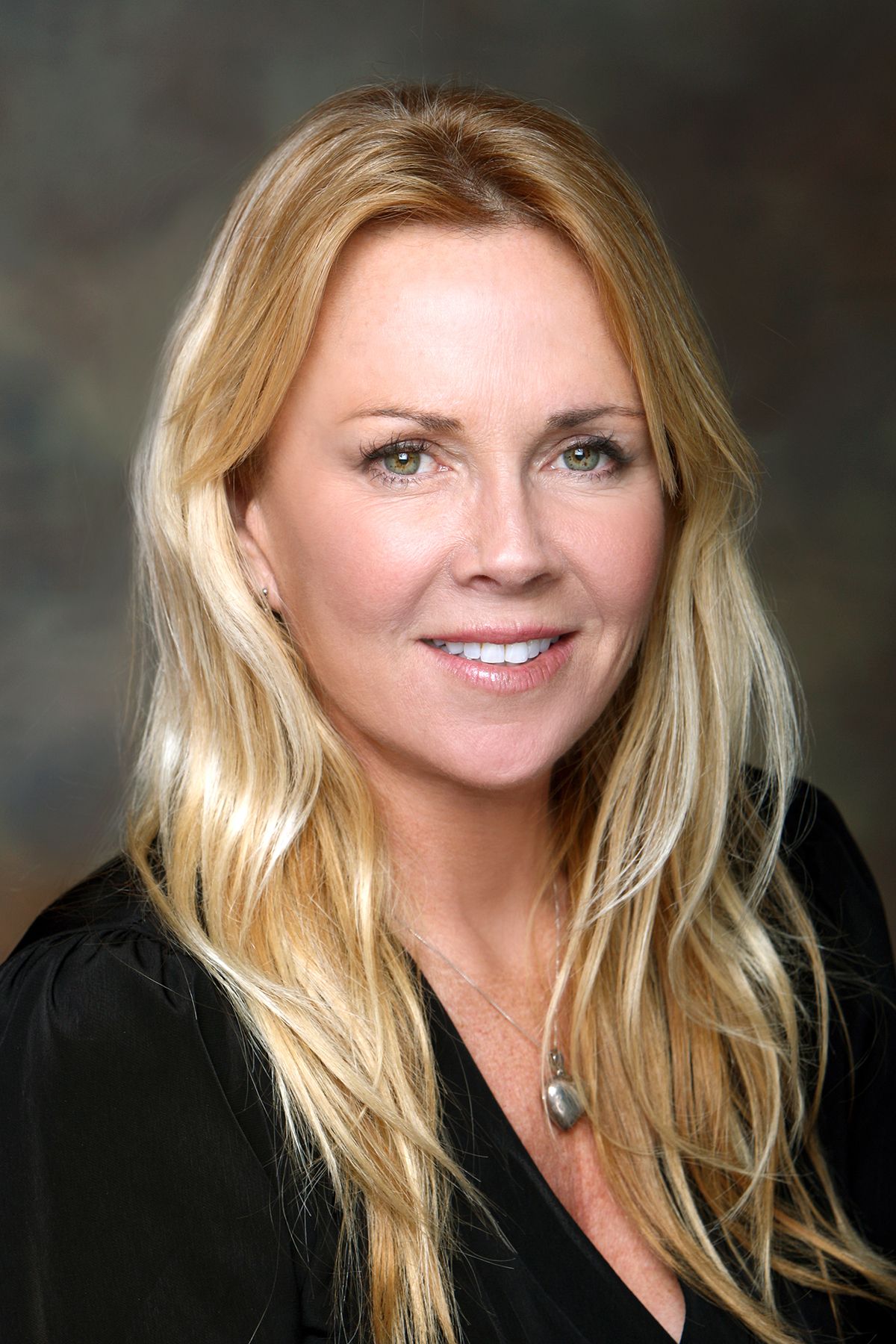 Professional headshot of a smiling blonde woman with long wavy hair wearing a black top and necklace.