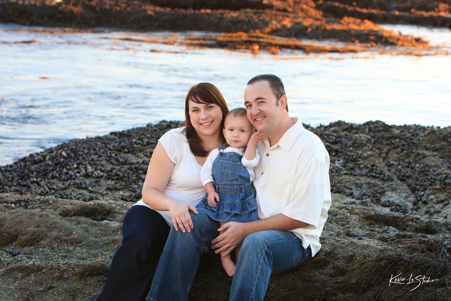 Young couple with toddler sitting together on rocky coastal shoreline.