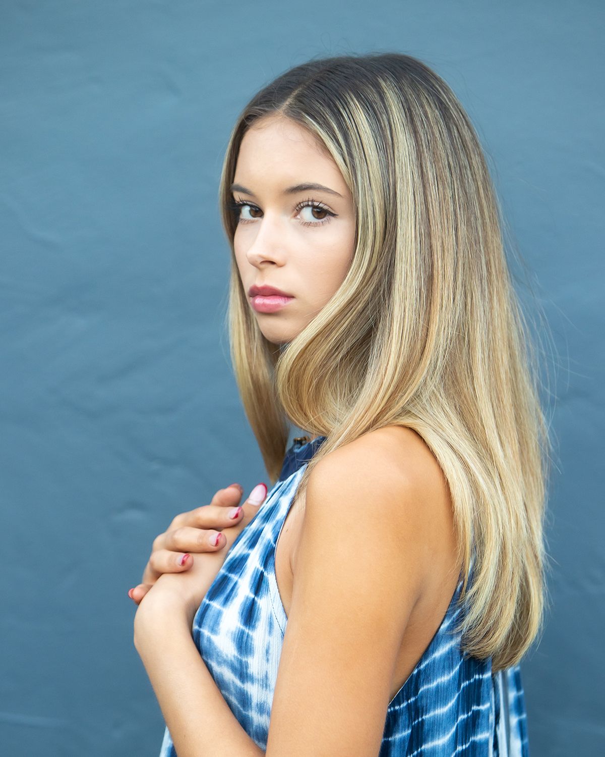 Young blonde woman in blue tie-dye top posing against a gray background.