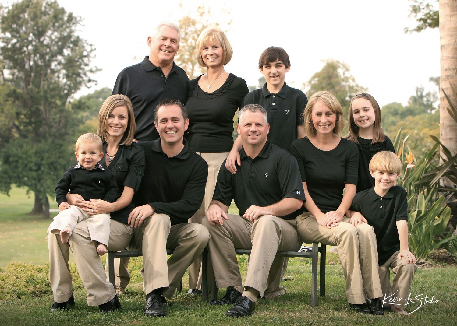 A large family poses outdoors on a sunny day, dressed in matching black tops and khaki pants for a professional portrait.