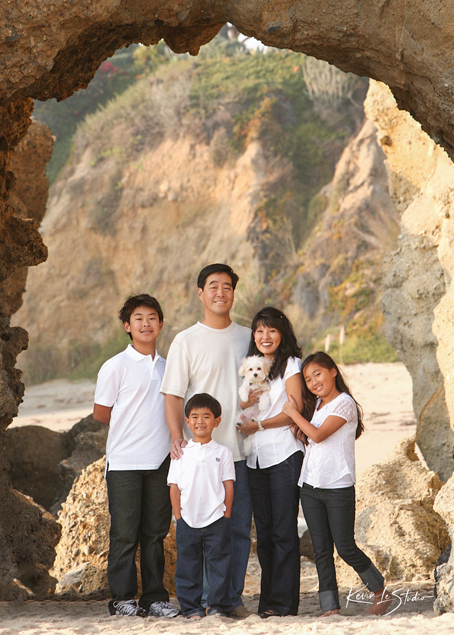 Asian family of four with small white dog posing under rock arch on sandy beach with cliffs in background.