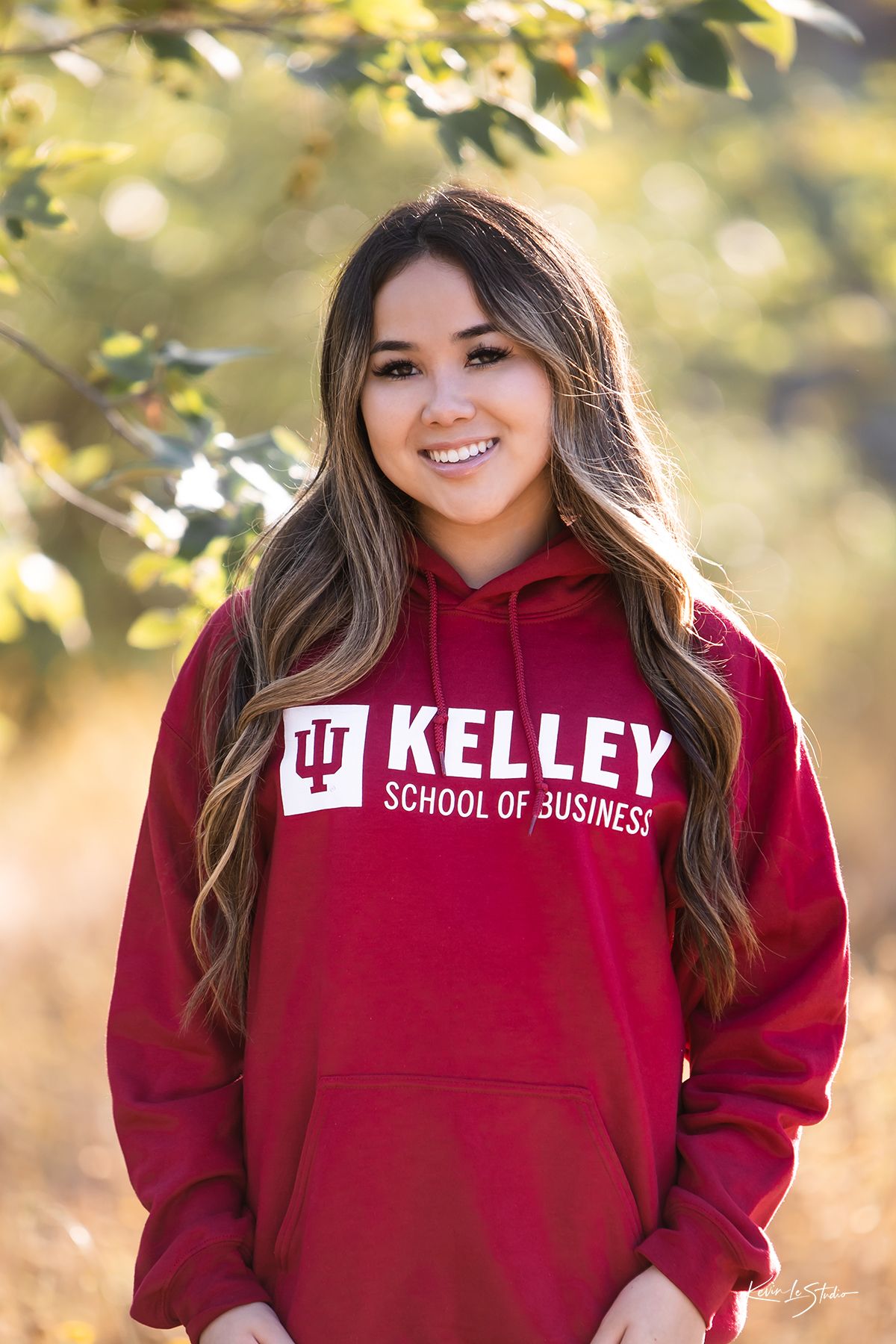 Smiling senior girl wearing Indiana University Kelley School of Business red hoodie poses outdoors in autumn setting.