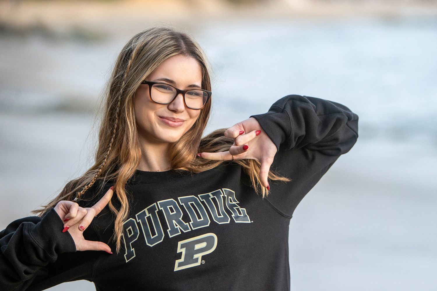 Smiling blonde girl in Purdue sweatshirt making hand gestures at the beach.