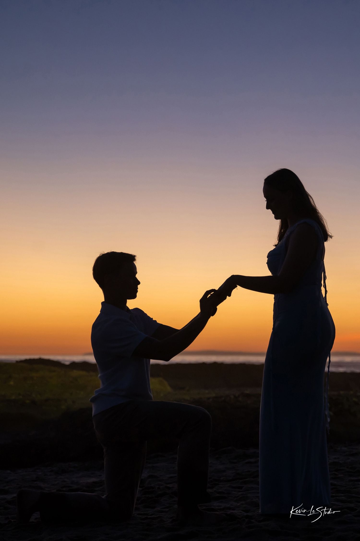 Silhouette of a man proposing on one knee to a woman at sunset on a beach with glowing orange sky.