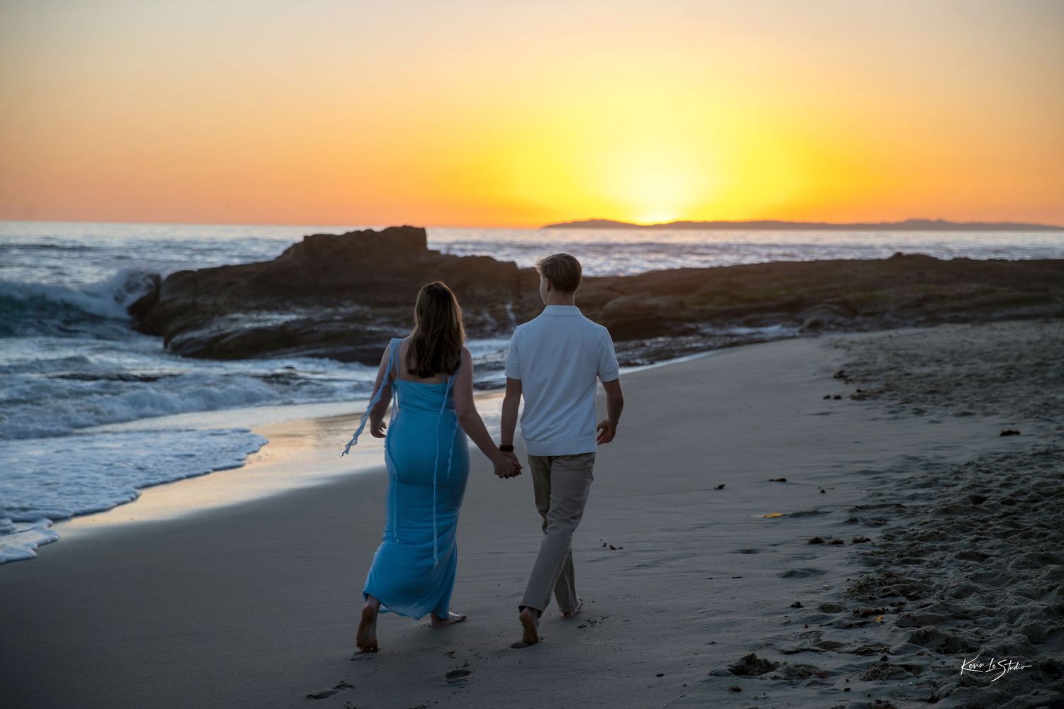 Couple walking hand in hand along sandy beach toward a vibrant orange and yellow sunset.