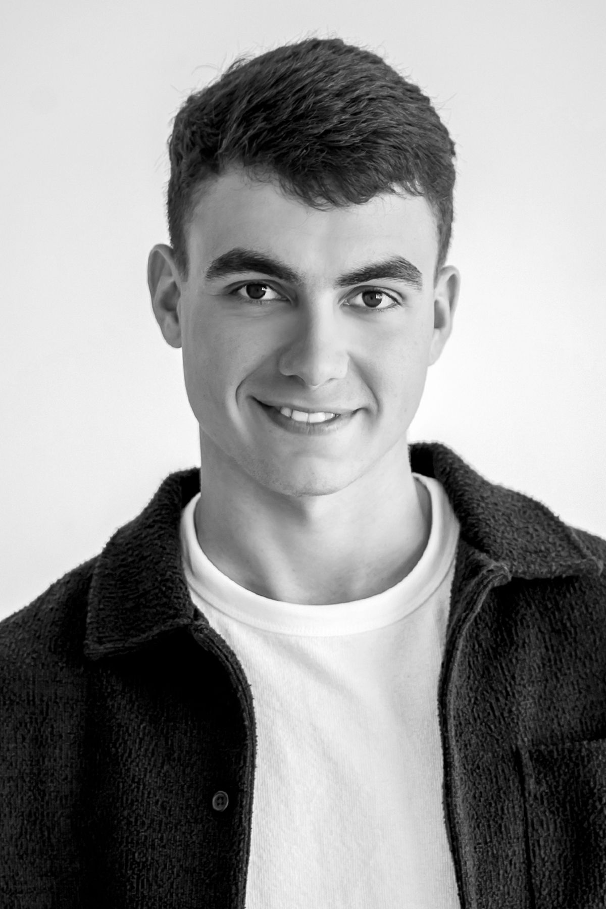 Black and white headshot of a smiling young man wearing a fluffy dark jacket over a white shirt.