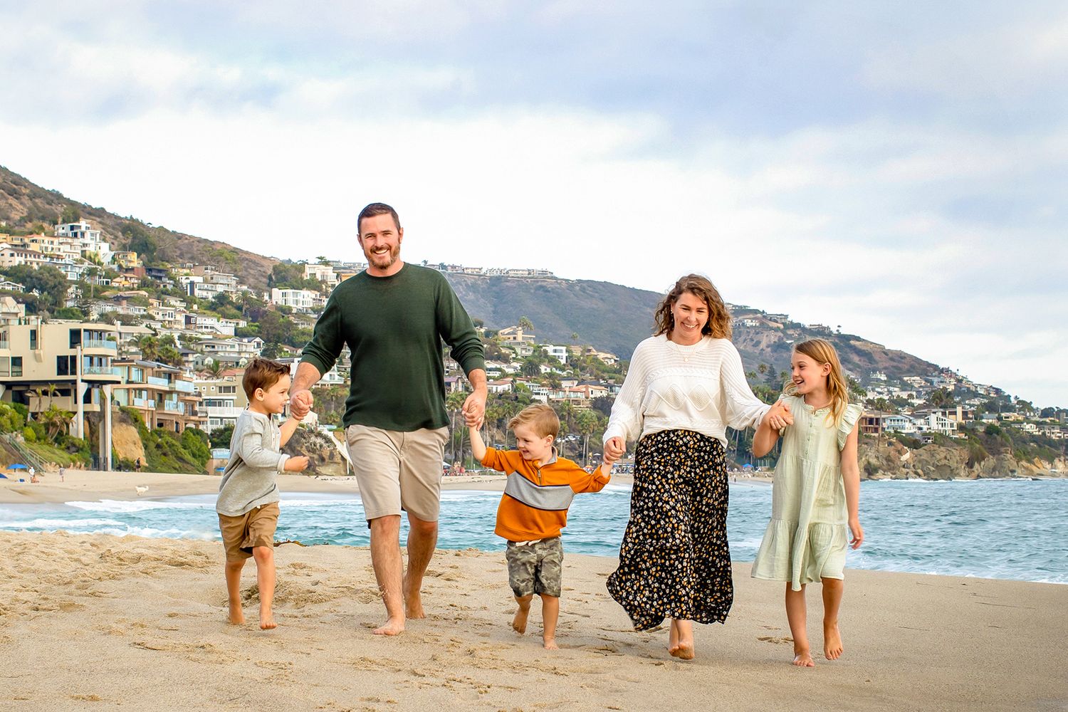 Happy family of five walking hand-in-hand on a sunny beach with hillside homes in the background.