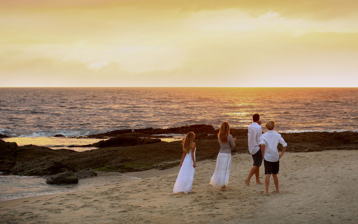 Family of four in white attire watching a golden sunset over the ocean on a rocky beach.