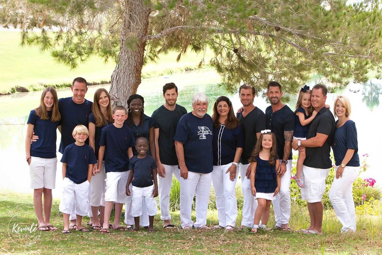 Large family group portrait outdoors near a lake, wearing coordinated navy and white outfits, smiling under a shady tree.