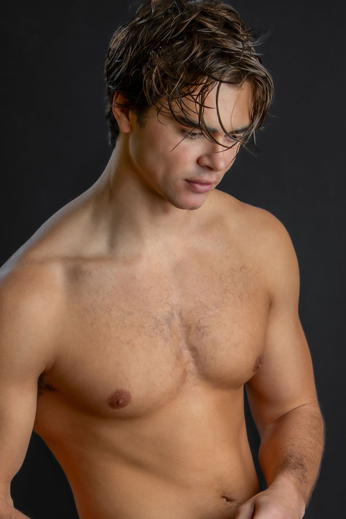 Shirtless athletic young man with wet brown hair posing against dark gray background in studio photo.