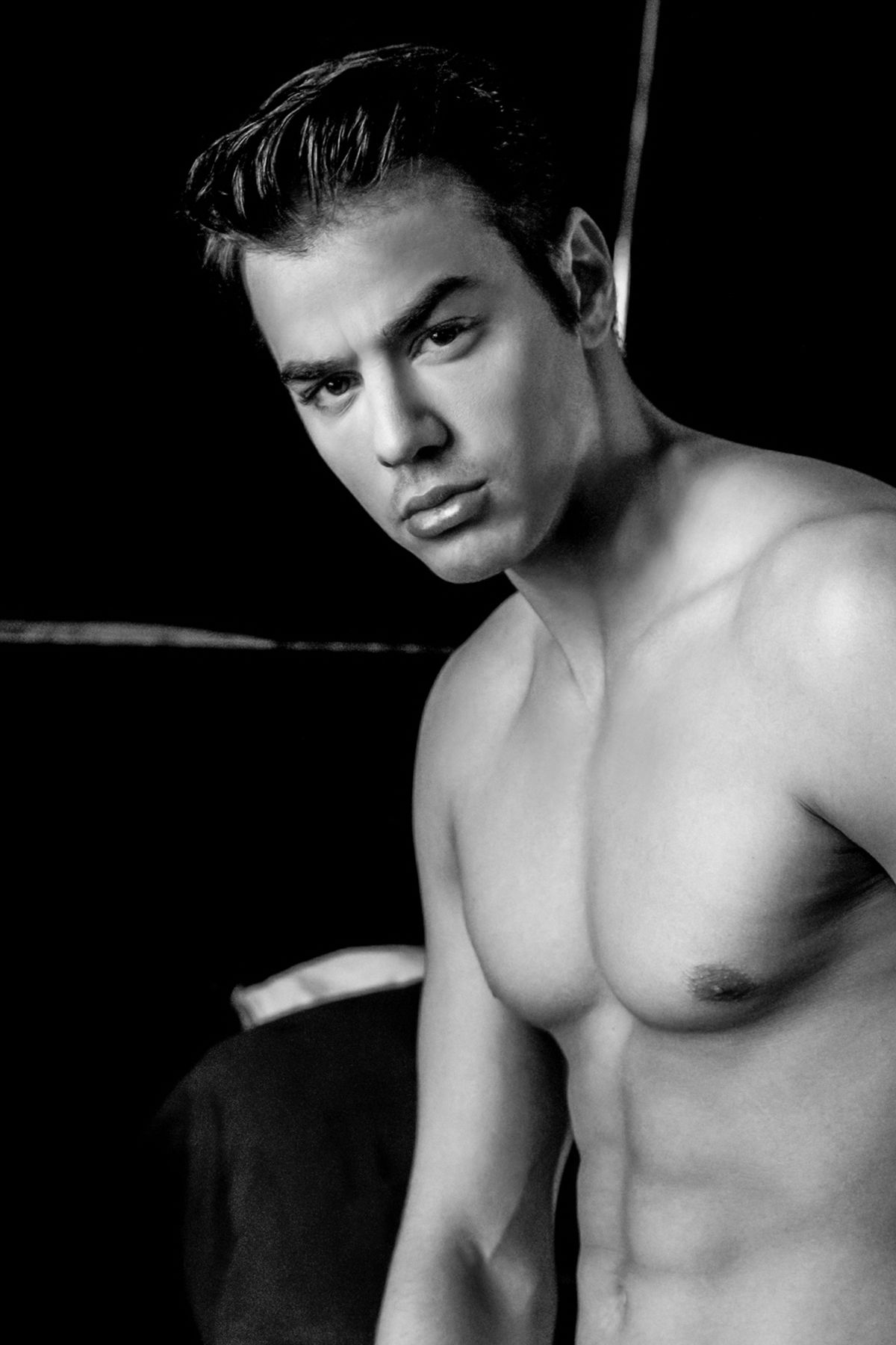 Black and white portrait of a shirtless muscular young man with styled hair posing against dark background.